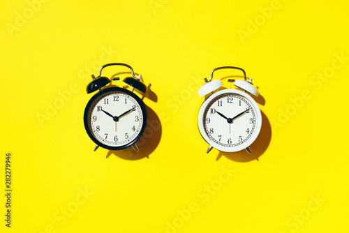 Black alarm clock and white one with hard shadow on yellow background. Top view
