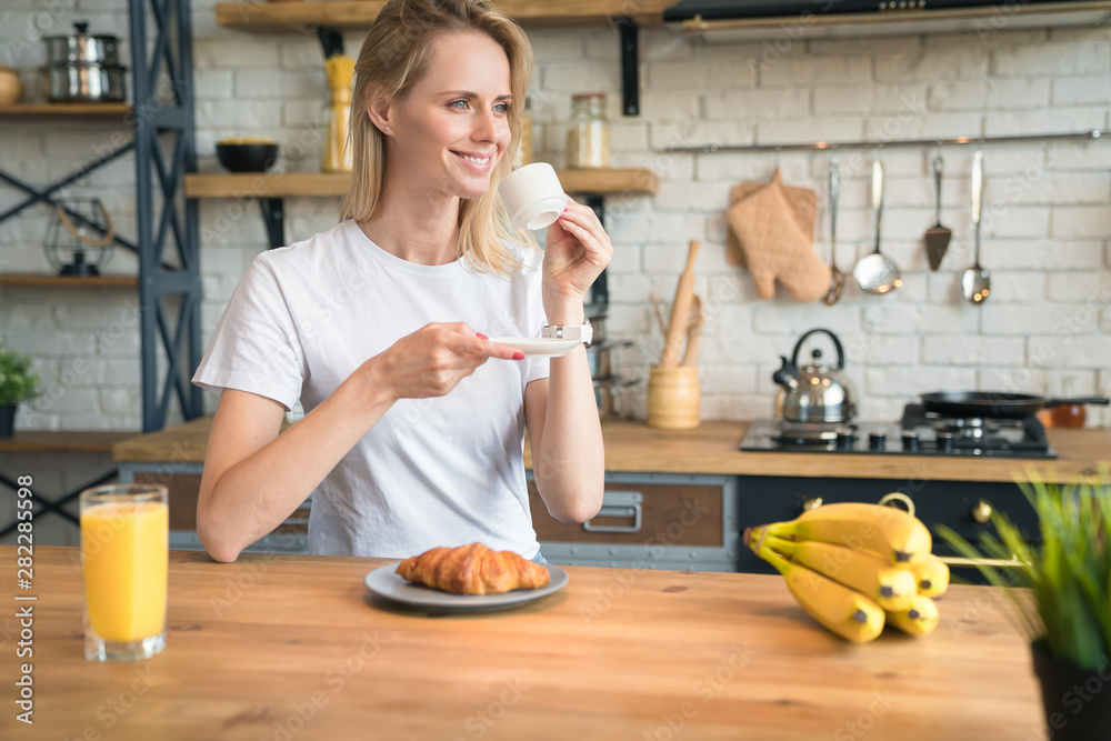pretty young smiling woman is sitting in the kitchen at home, having breakfast, drinking coffee with croissants and looking sideways. Morning home, Breakfast. wearing white shirt