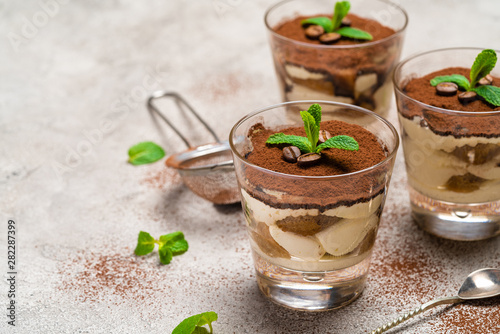 Classic tiramisu dessert in a glass cup and strainer with cocoa powder on concrete background