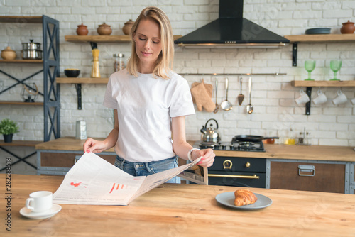young beautiful woman is reading her morning press while having breakfast in the kitchen. coffee with croissants. wearing white shirt and jeans