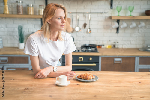 young beautiful woman is reading her morning press while having breakfast in the kitchen. coffee with croissants. wearing white shirt and jeans