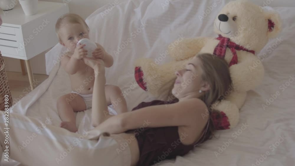 Beautiful young woman feeding her baby girl from the baby bottle lying in the bed. Concept of a happy family, one child, motherhood, love. Slow motion Beautiful young woman feeding her baby girl from the baby bottle lying in the bed. Concept of a happy family, one child, motherhood, love. Slow motion