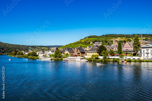 View to Middle Moselle River with cruise ships and Traben -part of the beautiful town of Traben-Trarbach with steeply sloped vineyards in the background, cloudless sky, Germany
