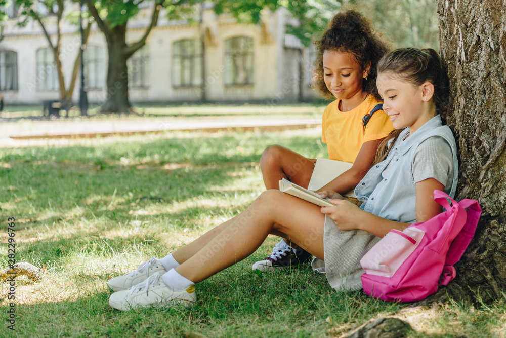 two cute multicultural schoolgirls sitting on lawn under tree and reading book together