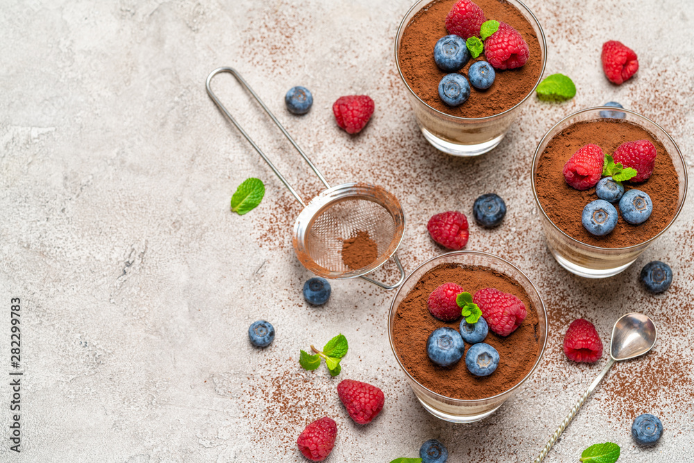 Classic tiramisu dessert with blueberries and raspberries in a glass and strainer with cocoa powder on concrete background