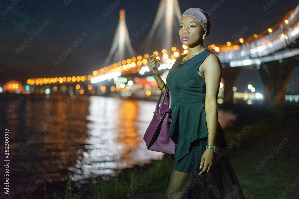Fototapeta premium African fashion model by the water. Girl in a black dress. Black girl in the night city. African American on the waterfront. A woman looks into the distance.Girl is waiting for the return of boyfriend