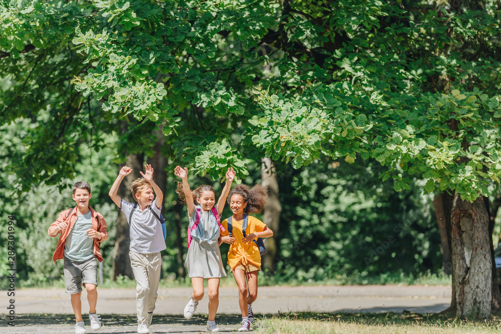 four happy multicultural schoolkids gesturing with raised hands while running in park four happy multicultural schoolkids gesturing with raised hands while running in park