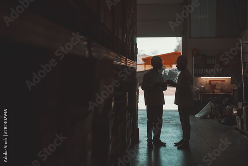 Silhouette of two hardworking warehouse workers standing in warehouse and checking inventory. Older one holding tablet while younger one looking at it.