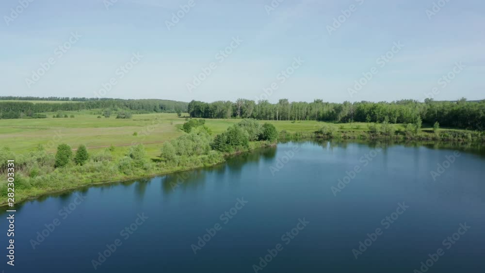 Aerial shot over the lake. Flying over a smooth surface of the water. View of the river bank with trees. In the distance are meadows and forests.