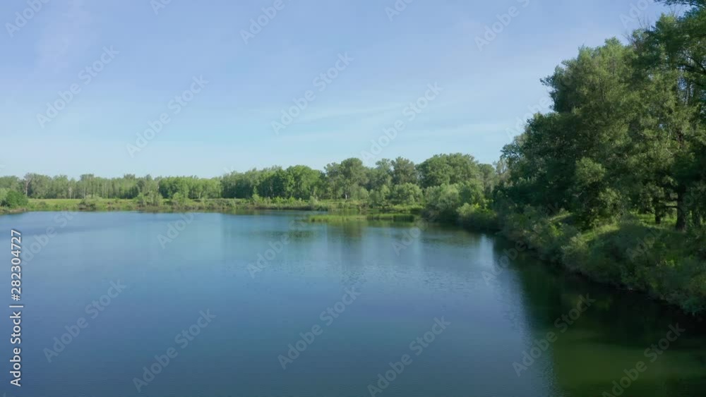 Aerial shot above the surface of the lake. Flying a drone along the riverbank with trees. On the water, you can also see a small island with plants. Aerial shot above the surface of the lake. Flying a drone along the riverbank with trees. On the water, you can also see a small island with plants.