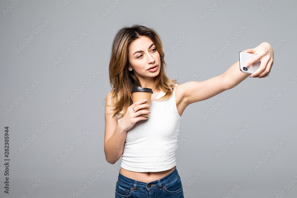 Portrait of happy woman taking selfie photo on smartphone in office and drinking takeaway coffee from plastic cup isolated over gray background - obrazy, fototapety, plakaty 