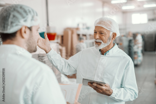 Two supervisors discussing about quality of food in food plant. Younger one holding folder with data while older one holding tablet. Both are dressed in white sterile uniforms and having hairnet.