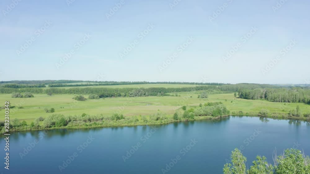 The descent shot over the lake shore. In the background is a view of green meadows and small sections of trees. Close up of trees on the shore. The descent shot over the lake shore. In the background is a view of green meadows and small sections of trees. Close up of trees on the shore.