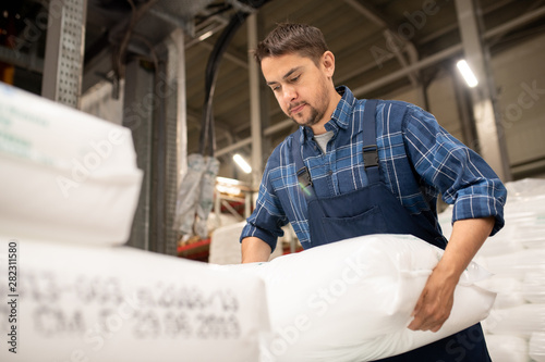 Young worker of polymer production factory loading sacks with plastic granules