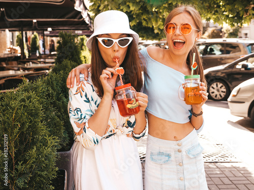 Two young beautiful smiling hipster girls in trendy summer clothes and panama hat.Sexy carefree women posing in the street.Positive models having fun in sunglasses.Drinking fresh cocktail smoozy drink