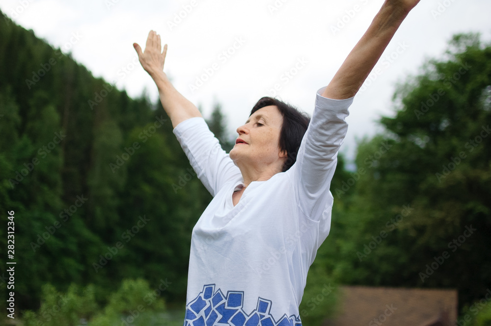 Senior woman doing a stretching exercise for the upper arms outside over landscape of forest and mountains Senior woman doing a stretching exercise for the upper arms outside over landscape of forest and mountains