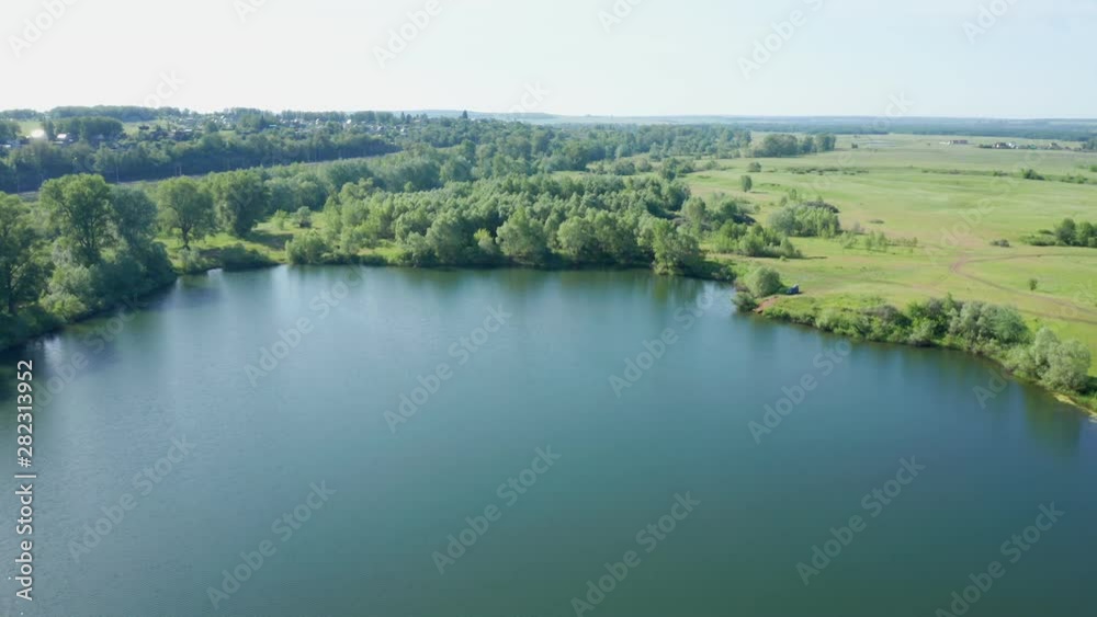 Aerial shot over the lake on a sunny day. View of the lake with dense vegetation. There is a car on the shore. In the distance is a village. Aerial shot over the lake on a sunny day. View of the lake with dense vegetation. There is a car on the shore. In the distance is a village.