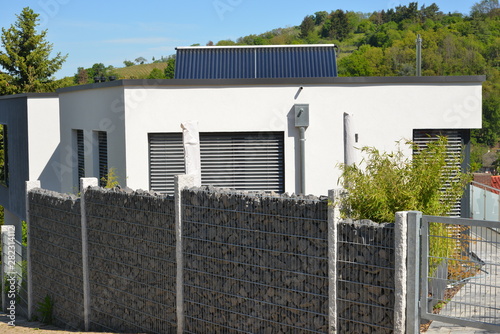 Moderner Sichtschutzzaun aus mit Naturstein gefüllten Gabionen zwischen Granit-Säulen, vor einem modernen Flachdach-Bungalow mit Solaranlage und integrierter Dachrinne