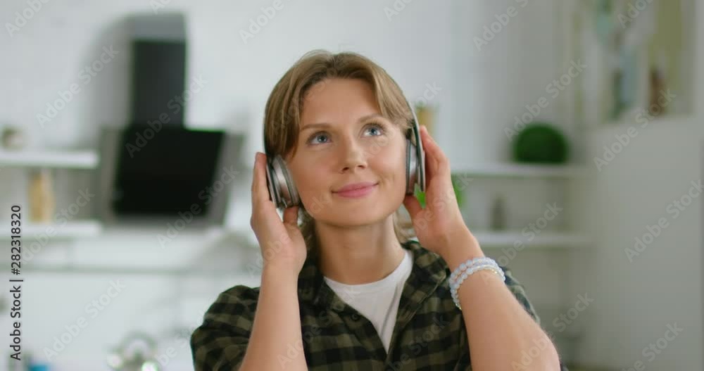 Young short-haired woman in casual shirt is listening music in wireless headset at home, relaxing, easy-listening, enjoying her freetime, smiling, being in a good mood, indoor. 4K, shot on RED camera. Young short-haired woman in casual shirt is listening music in wireless headset at home, relaxing, easy-listening, enjoying her freetime, smiling, being in a good mood, indoor. 4K, shot on RED camera.