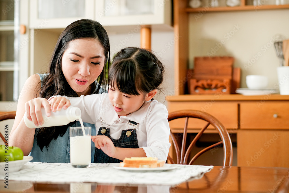 Asian mother take care her daughter pour milk to a glass on the table in house kitchen during breakfast meal. Main focus is the mother. Asian mother take care her daughter pour milk to a glass on the table in house kitchen during breakfast meal. Main focus is the mother.