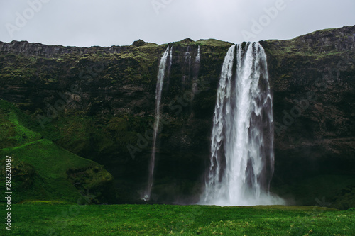 Famous Seljalandsfoss waterfall in Iceland. Saturated green grass  pale gray rocks  and heavy rainy sky