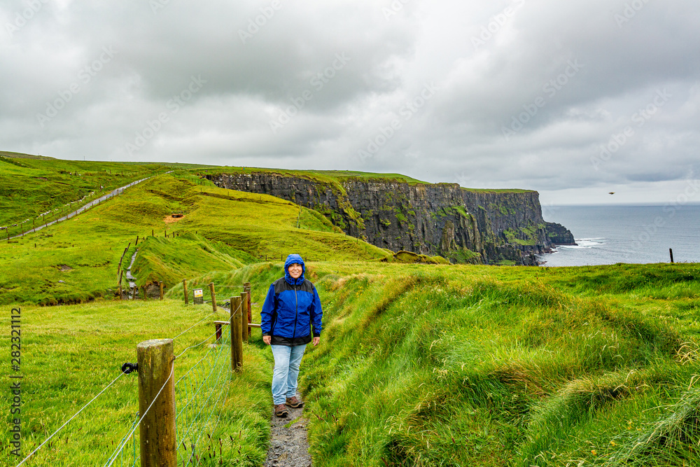Woman in blue jacket walking the coastal walk route from Doolin to the Cliffs of Moher, geosites and geopark, Wild Atlantic Way, spring rainy day in the countryside in county Clare in Ireland