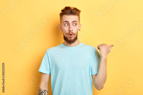 emotional funny man looking aside, being horrified to see something, indicates with thumb aside, isolated over yellow background. close up portrait. copy space.place for text. look here