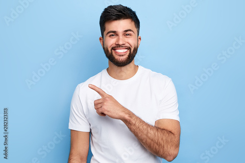 Young handsome charming man showing something with index finger, place for text, advert, copy space. isolated on blue background. direction. body language. sale, shop photo