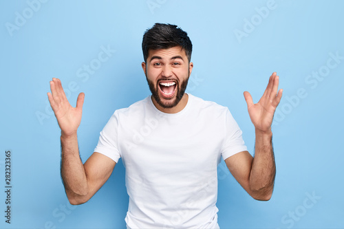 Cheerful handsome happy guy with wide opened mouth raising his hands up, having eyes full of happiness, rejoicing his achievements.