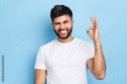 happy funny bearded man winking his eye and showing ok sign, isolated on blue background, everything is under control. joke , fun concept