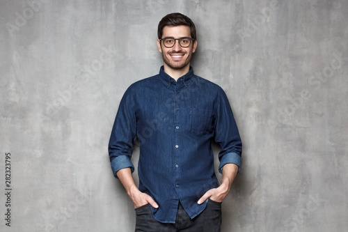 Young smiling man wearing trendy glasses and denim shirt, leaning on gray textured wall