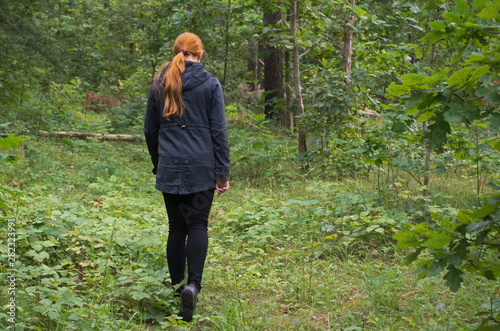 Redhaired girl in a black jacket and leggings is on the green grass of the forest among the trees © ANDY RELY