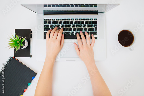 Top view of female hands typing on a laptop keyboard. Coffee, plant and notebook beside.