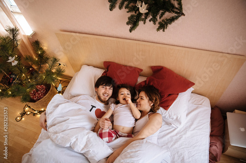 Family portrait of mother, father and daughter in white clothes having fun on the bed and christmas tree near them
