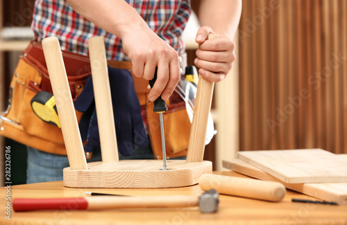 Fototapeta Young working man repairing wooden stool using screwdriver indoors, closeup