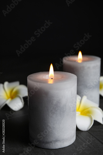 Burning candles and plumeria flowers on dark grey table against black background  space for text
