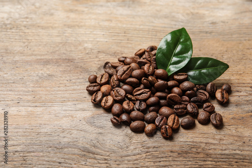 Pile of coffee beans and fresh green leaves on wooden table, space for text