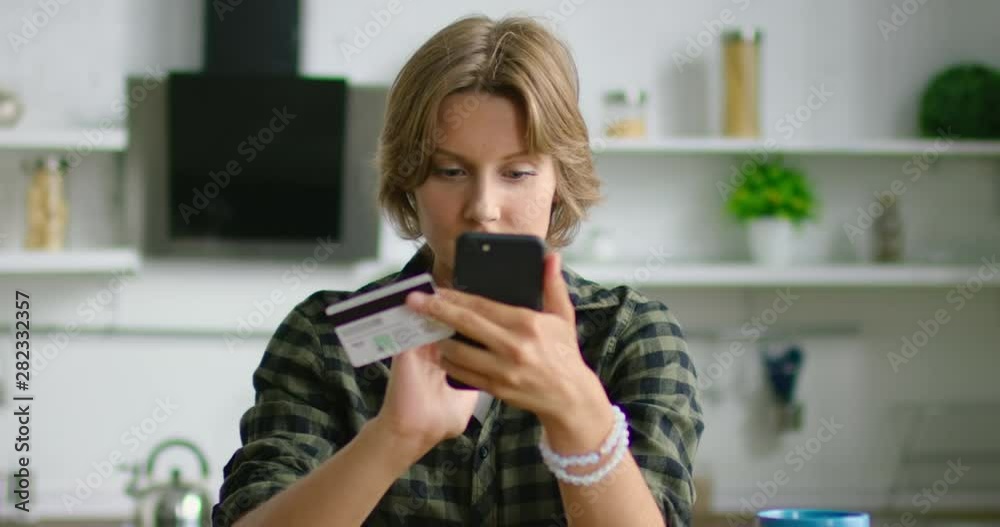 Young woman is entering credit card number in her smartphone, gadget, doing online shopping, happy and cheerful, sitting at table in kitchen, Caucasian, white. 4K, shot on RED camera. Young woman is entering credit card number in her smartphone, gadget, doing online shopping, happy and cheerful, sitting at table in kitchen, Caucasian, white. 4K, shot on RED camera.