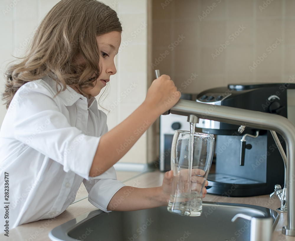 Portrait of a little caucasian girl gaining a glass of tap clean water. Kitchen faucet. Cute curly kid pouring fresh water from filter tap. Indoors. Healthy life concept Portrait of a little caucasian girl gaining a glass of tap clean water. Kitchen faucet. Cute curly kid pouring fresh water from filter tap. Indoors. Healthy life concept