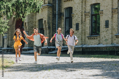 four adorable multicultural schoolchildren smiling while running in schoolyard