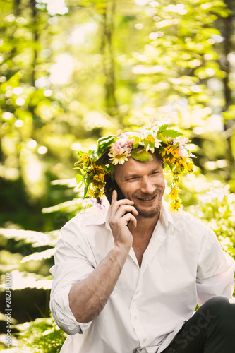 close-up portrait of a young handsome guy hipster groom dressed in stylish white shirt and a wedding wreath of flowers on his head on a background of summer forest talking on phone