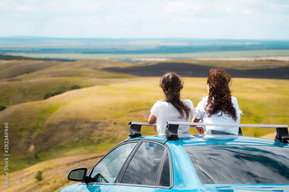 Two young girlfriends travel by car in the mountains. Two women are looking out of the car door at the nature around