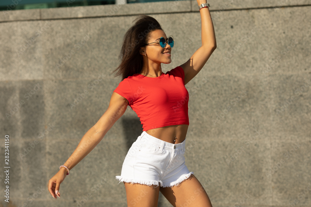 Fototapeta premium Portrait of trendy young black woman in white shorts and red top walking outdoors .