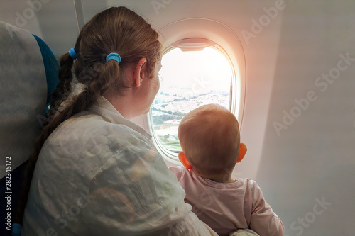 Mom with a small baby in her arms flying on the plane. They look out the window with interest on the earth. First time traveling with children