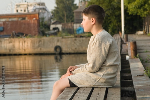 A boy in a linen shirt sits on the edge of a wooden pier in the summer and looks at the water © Sergey