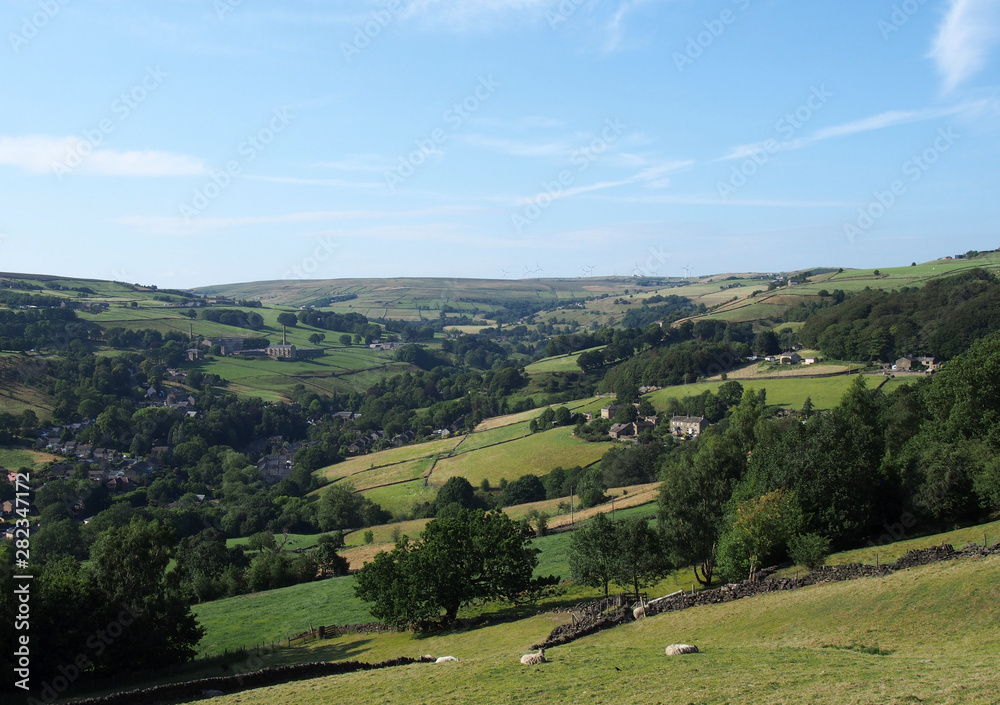 a panoramic view of the village of luddenden in west yorkshire surrounded by fields with grazing sheep and trees in summer sunlight