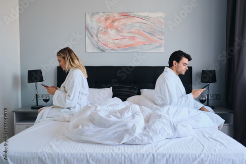 Young brunette man and pretty blond woman in white bathrobes using cellphones sitting on different sides of bed in modern hotel