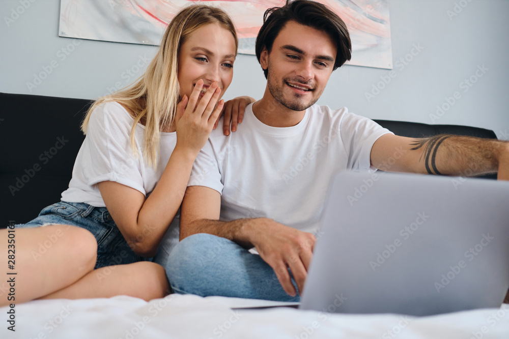 Beautiful blond woman and smiling brunette man happily watching movie on laptop together. Young beautiful couple in white T-shirts sitting on bed at home Beautiful blond woman and smiling brunette man happily watching movie on laptop together. Young beautiful couple in white T-shirts sitting on bed at home