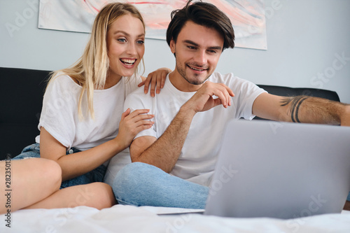Pretty emotional blond woman and attractive smiling brunette man happily watching movie on laptop together. Young beautiful couple in white T-shirts sitting on bed at home