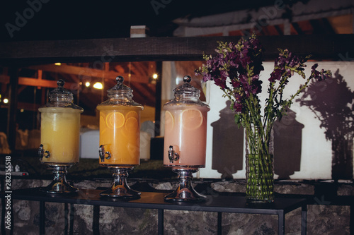 Three pitchers of orange, grapefruit and lemon juices, a glass vase with lilac flowers on black metal tables.